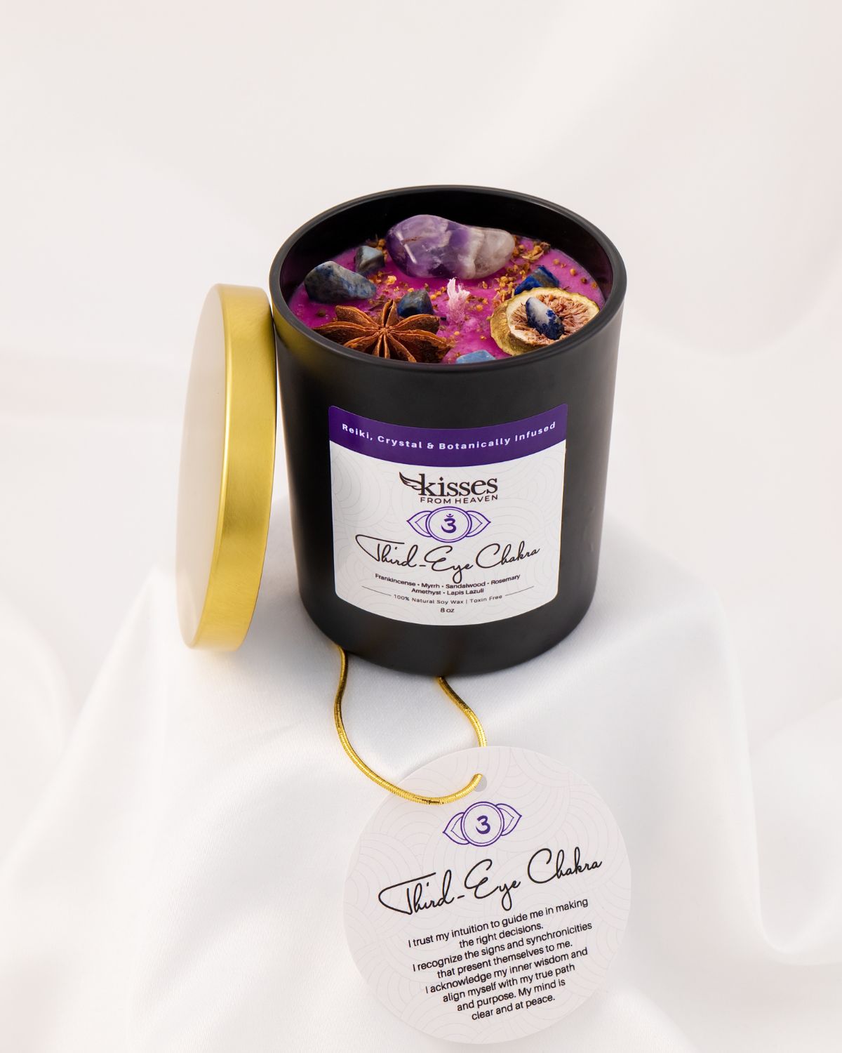 A black cylindrical candle jar with its gold lid leaning against it, filled with colorful wax and embedded crystals and dried botanicals. The front label reads 'Kisses' and 'Third Eye Chakra,' with a matching circular tag hanging below on a white draped fabric background.