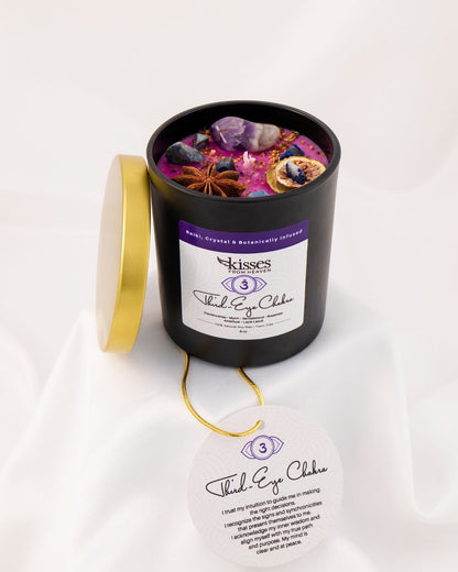 A black cylindrical candle jar with its gold lid leaning against it, filled with colorful wax and embedded crystals and dried botanicals. The front label reads 'Kisses' and 'Third Eye Chakra,' with a matching circular tag hanging below on a white draped fabric background.