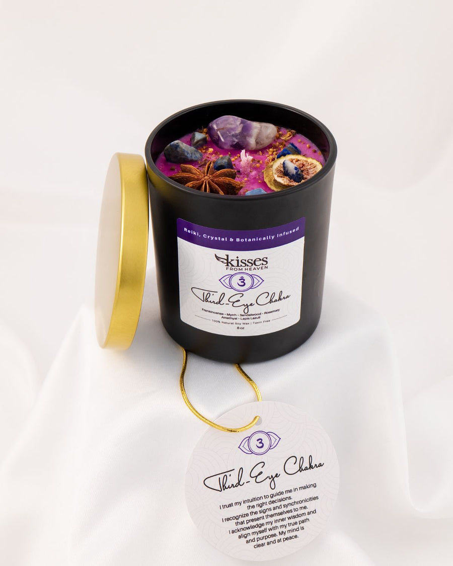 A black cylindrical candle jar with its gold lid leaning against it, filled with colorful wax and embedded crystals and dried botanicals. The front label reads 'Kisses' and 'Third Eye Chakra,' with a matching circular tag hanging below on a white draped fabric background.