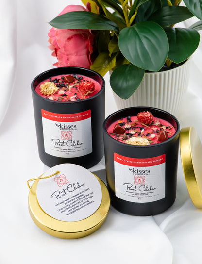 Two black candles with floral toppings on a white surface, surrounded by flowers and a plant.
