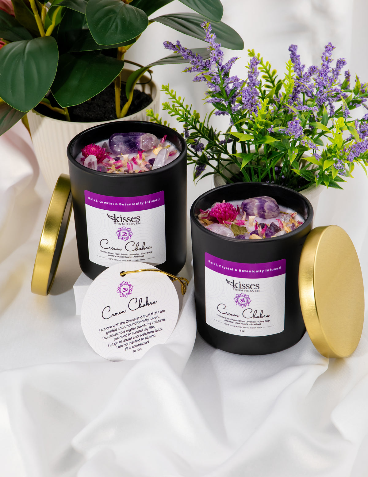 Two black candles with gold lids on a white fabric background, surrounded by plants.