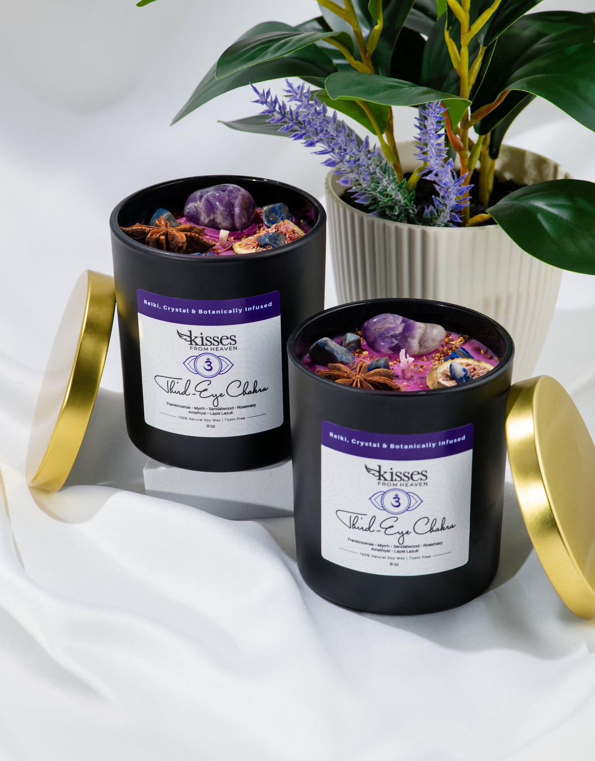 Two black candles with gold lids on a white surface, surrounded by plants.
