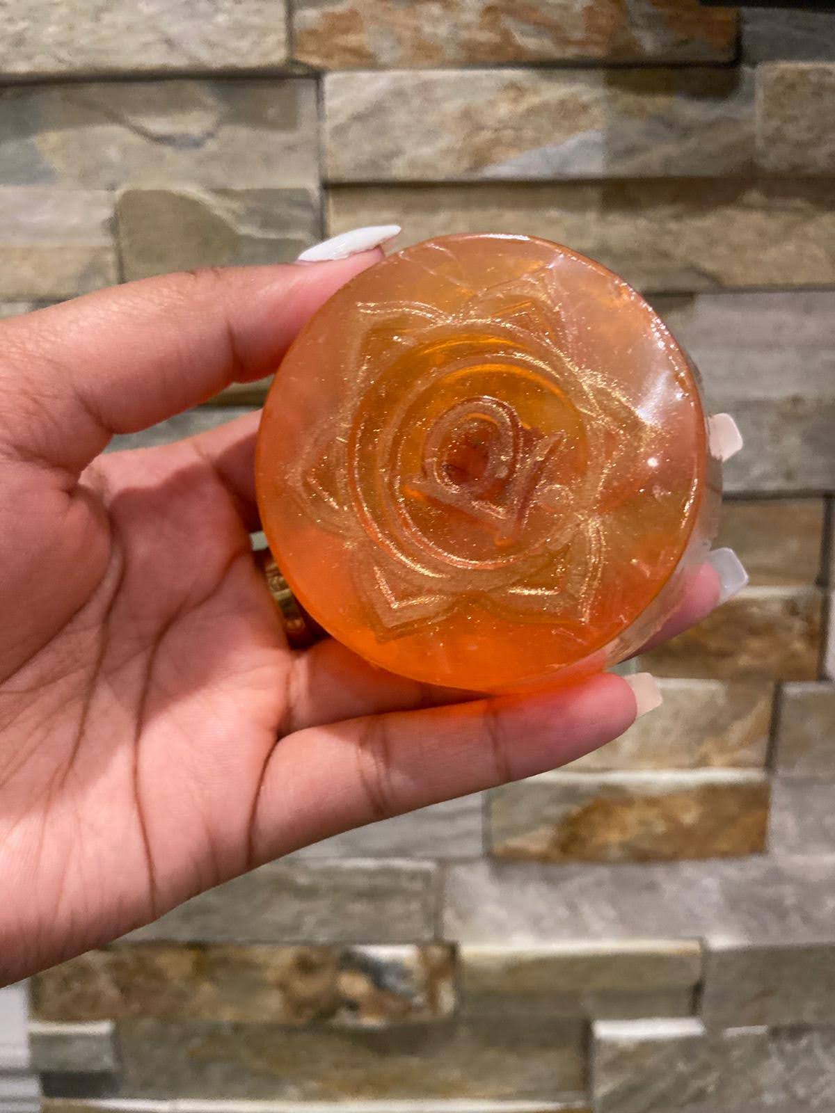 Hand holding a round, orange-colored soap with a rose design against a stone wall background